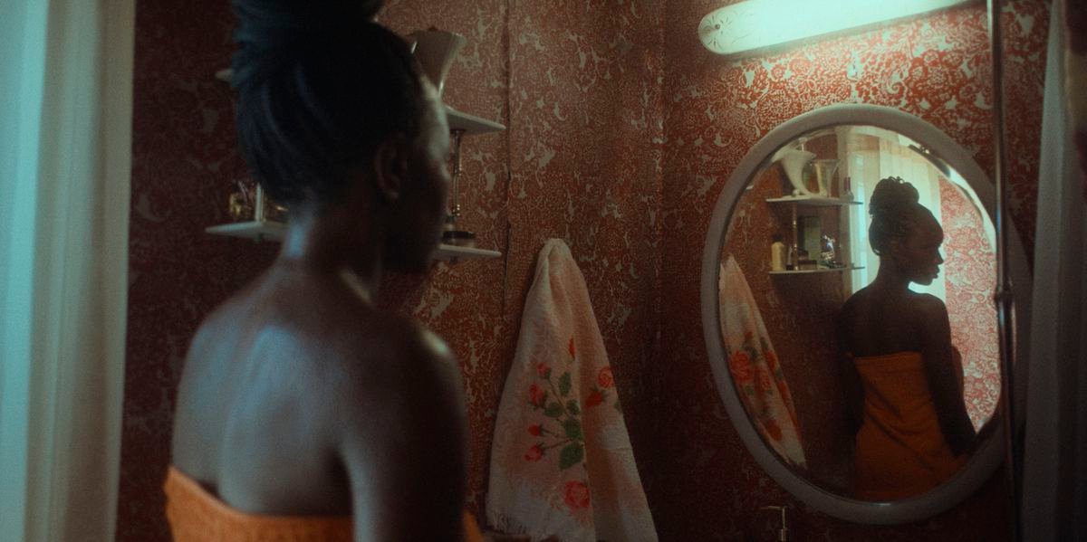 Aisha (Anna Diop), a dark-skinned woman wearing a bright orange towel, examines herself in a mirror in a darkened room in Nanny
