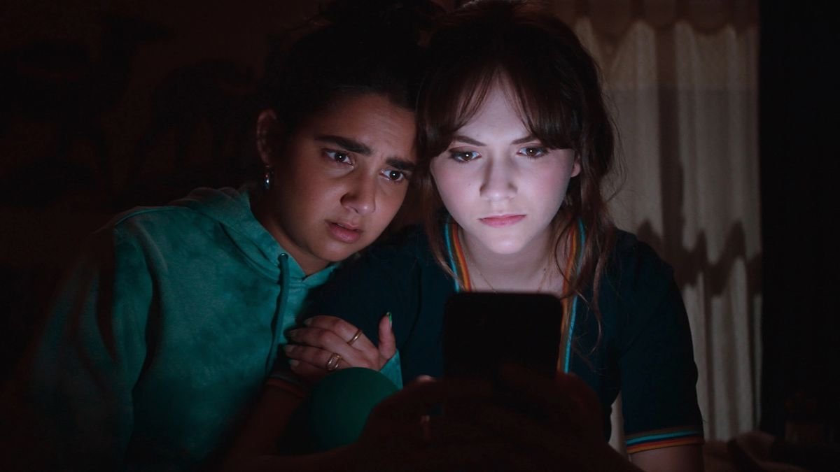 Cat Person stars Emilia Jones and Geraldine Viswanathan huddle around a mobile device, lit by the glow as they scroll