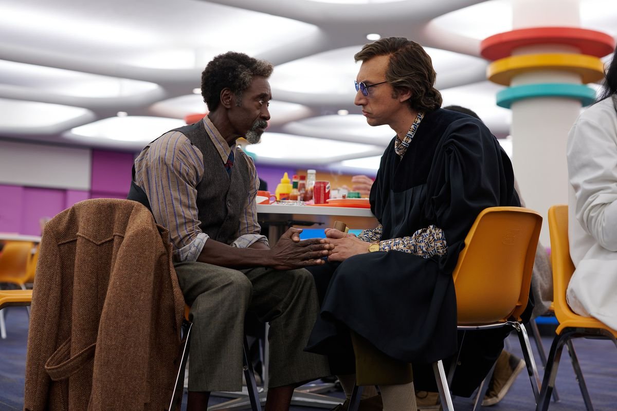 Adam Driver, wearing an academic gown and dark glasses, chats with Don Cheadle in a colorful, retro canteen