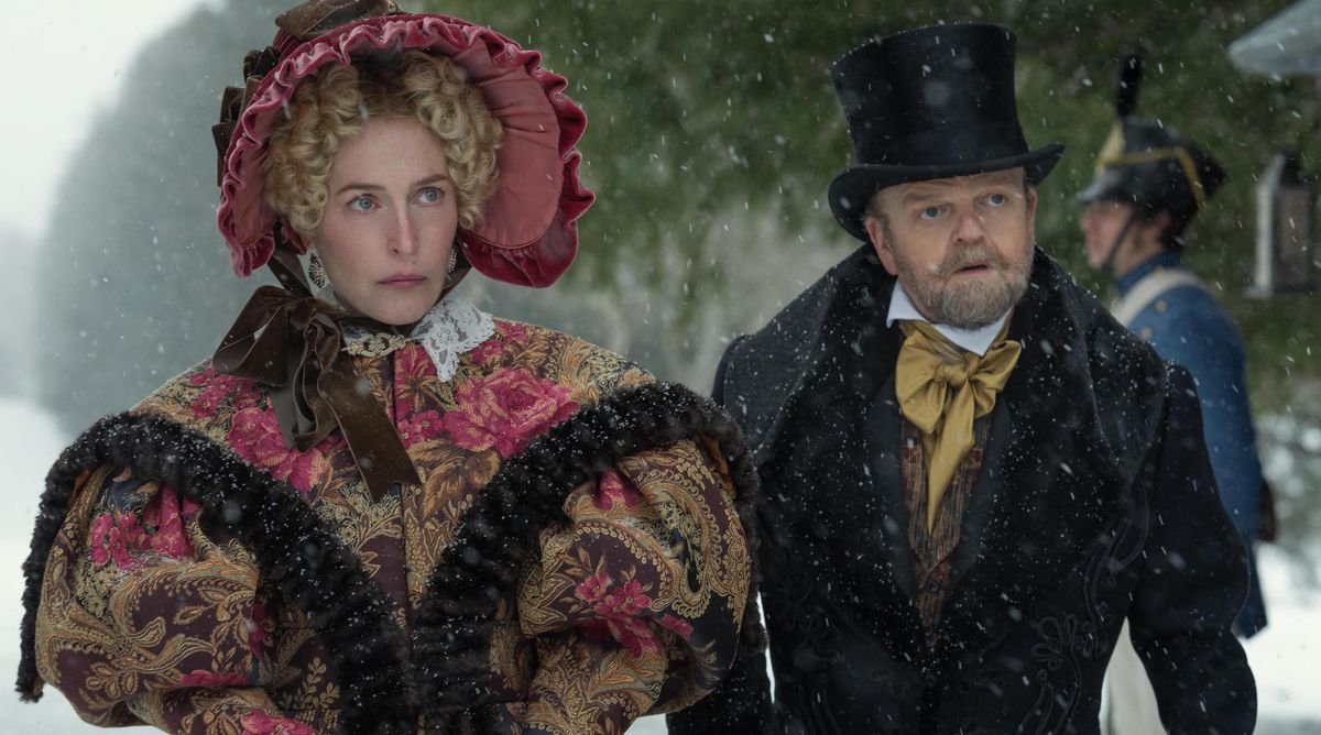 Gillian Anderson in an incredibly bulky brocade wrap and bonnet and Toby Jones in a top hat and suit stand in the snow as Julia and Dr. Marquis in The Pale Blue Eye