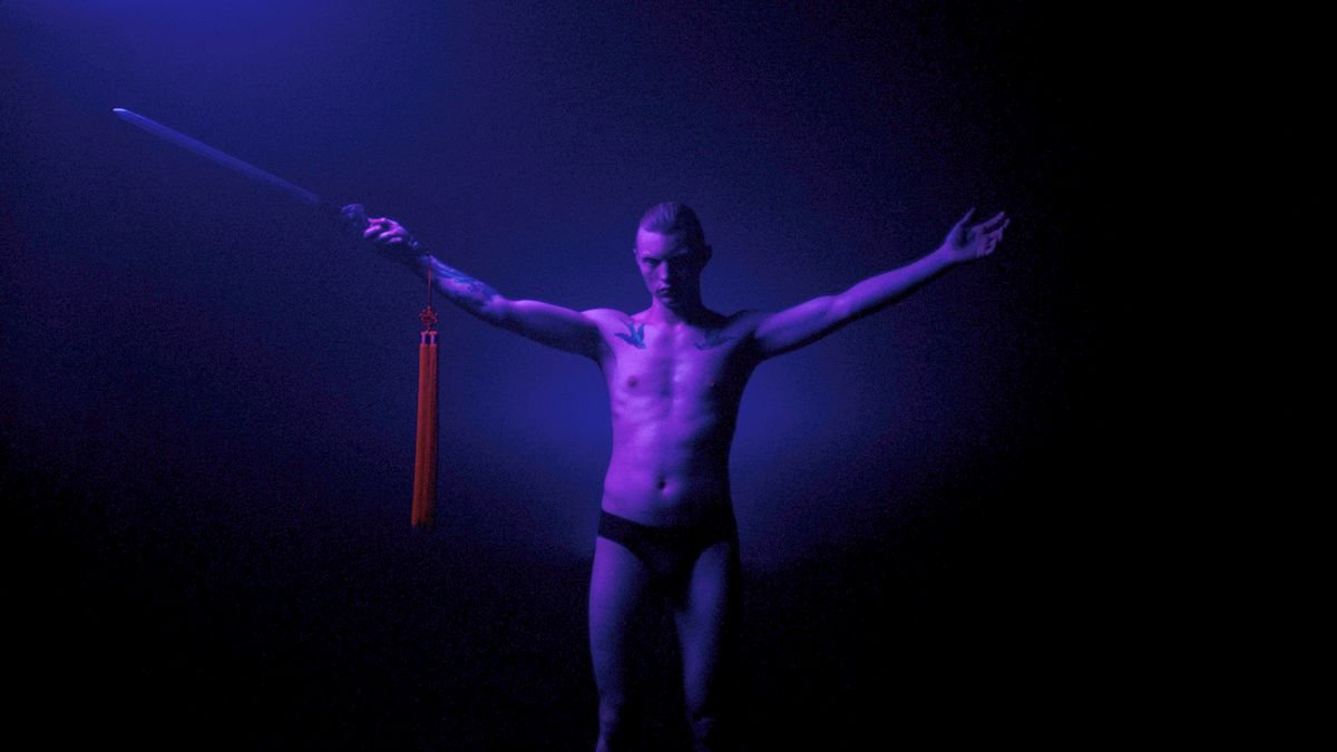 A man in Copenhagen Cowboy standing in purple neon light holding a sword with his arms in a Y position