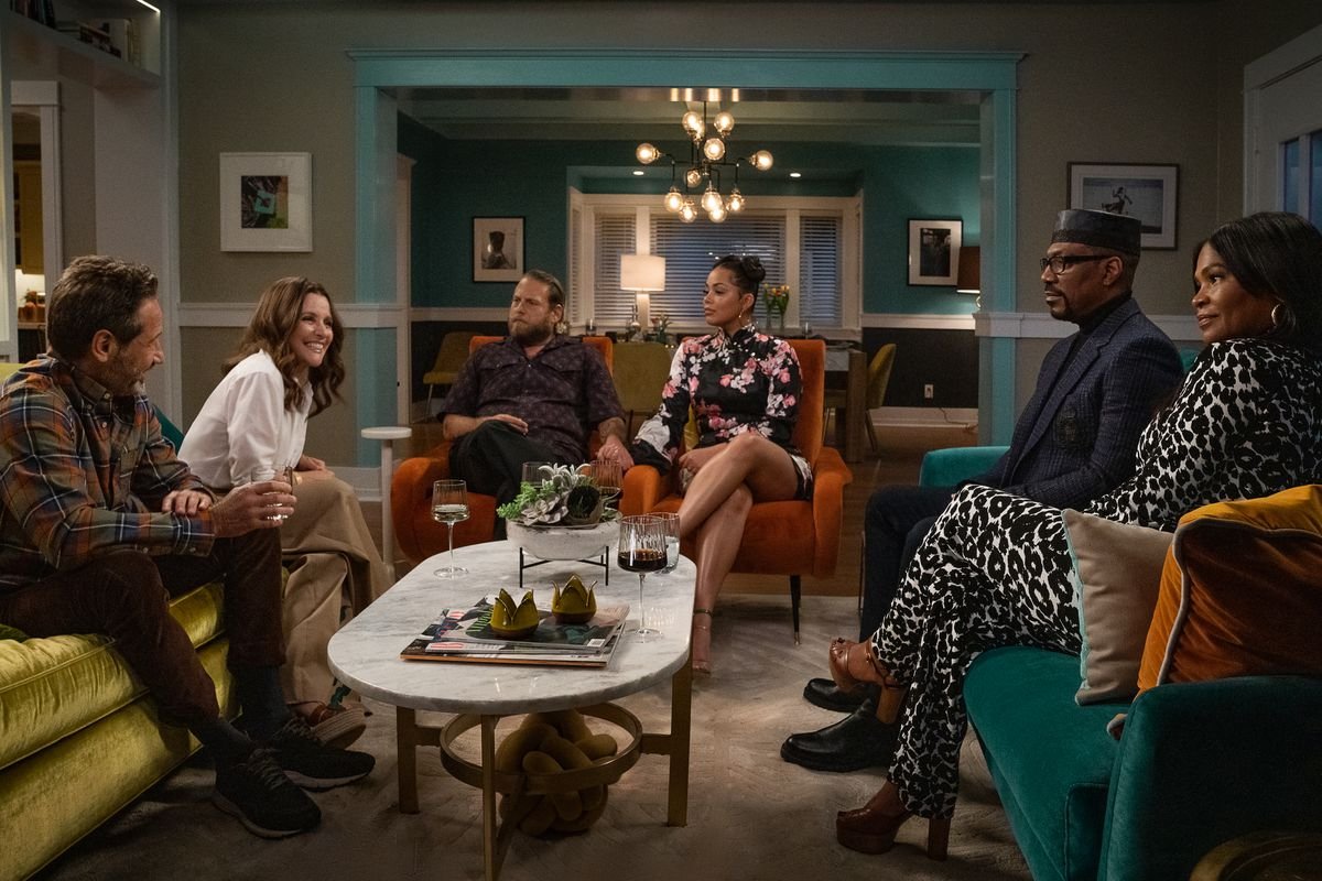 The Cohen and Muhammed families sit in a living room together in awkward politeness in a scene from Netflix’s You People. 