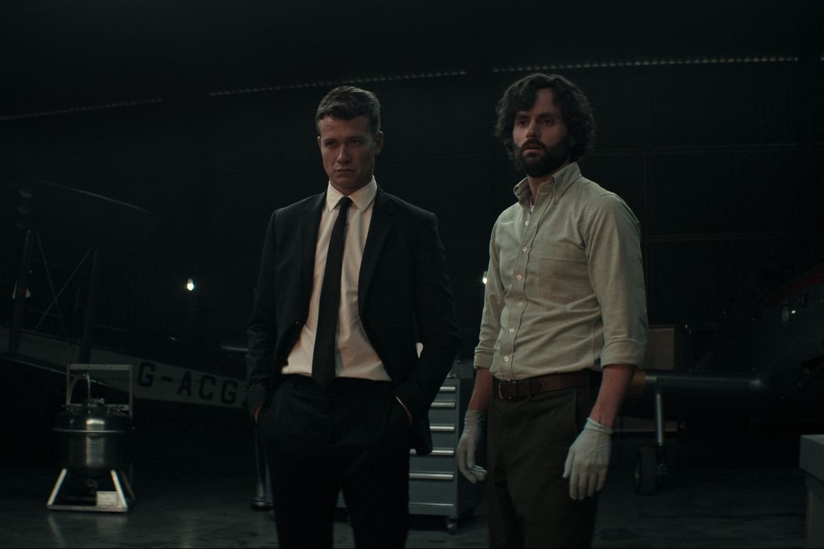 Rhys in a black suit, and Joe in a shirt with rolled up sleeves and latex gloves, stand next to each other in a dark basement in You season 4