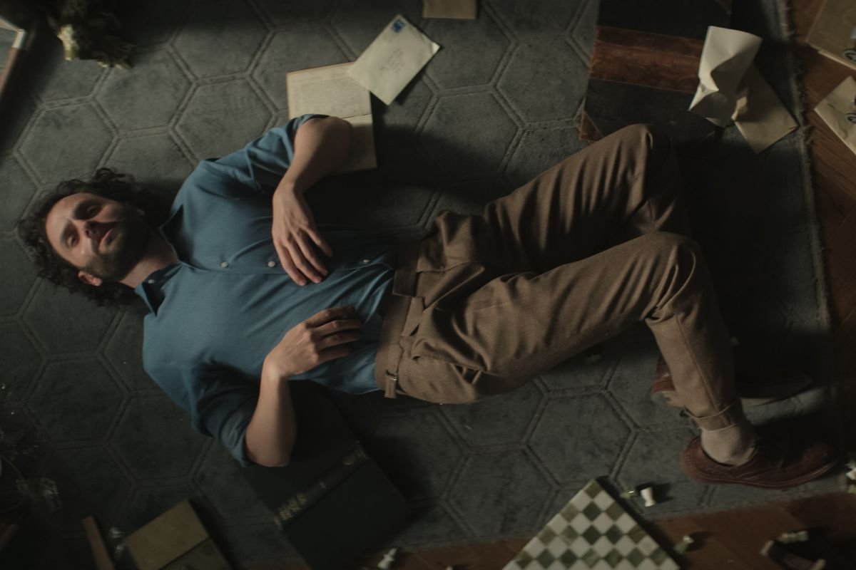 Joe Goldberg lies on the floor of his flat with books, papers, and a chessboard around him in You season 4.
