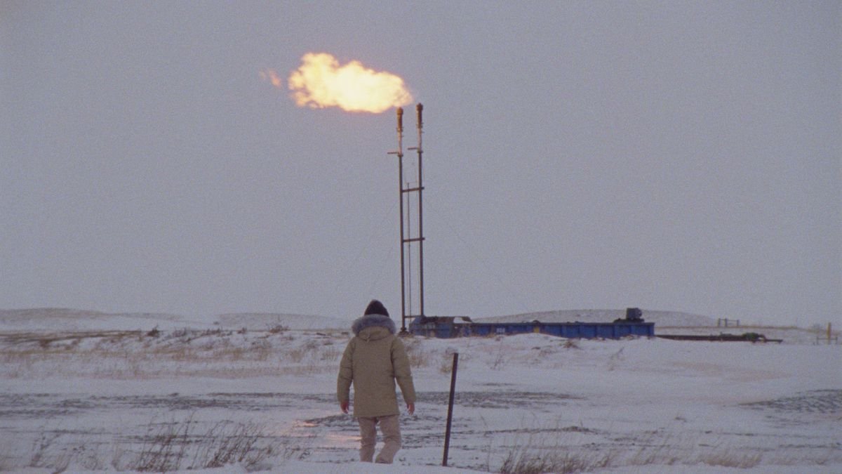 A man in a parka walks in the snow as flames roar from a smokestack ahead of him in How to Blow Up a Pipeline