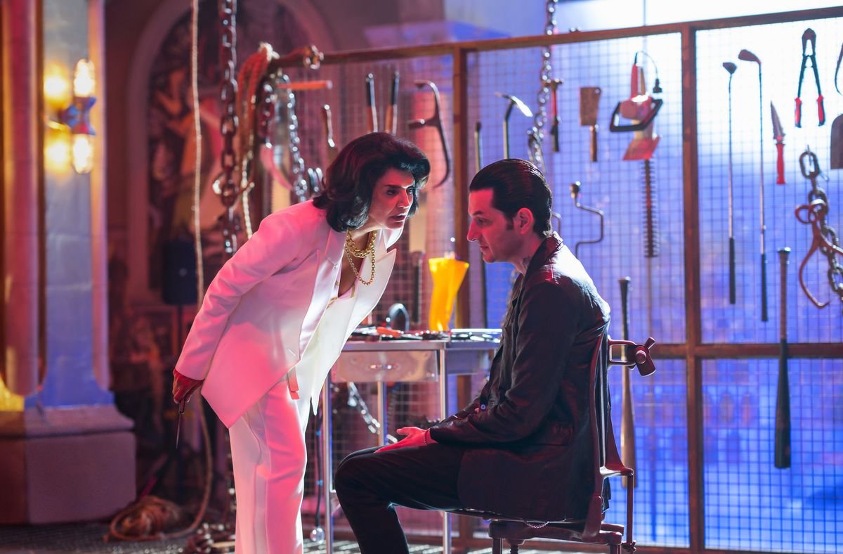 In a production shot from Renfield, Bellafrancesca Lobo (Shohreh Aghdashloo, in a white pantsuit and heavy gold chains) leans over a seated, black-suited Tedward Lobo (Benjamin Schwartz) in what appears to be a neon-lit torture chamber, with chains dangling from the ceiling and a backlit mesh-fence wall covered with garden tools, weapons, and random knives