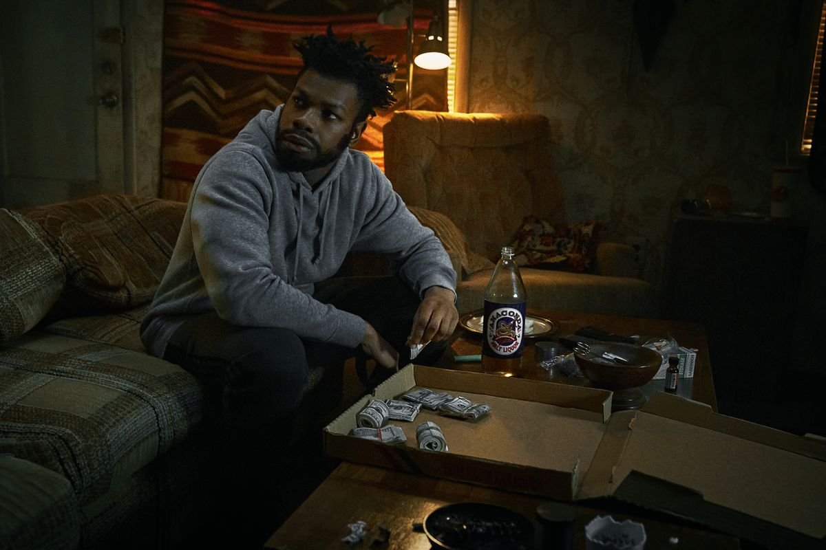 John Boyega as Fontaine sitting on a couch in front of a pizza box filled with rolls of dollar bills in They Cloned Tyrone.