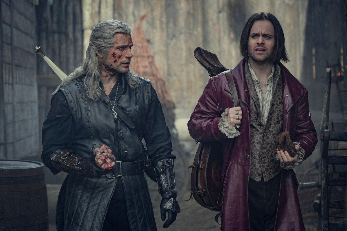 Geralt, bloody and carrying a monster heart after a battle, walks next to Jaskier the bard, who is carrying a coin purse and very perplexed.