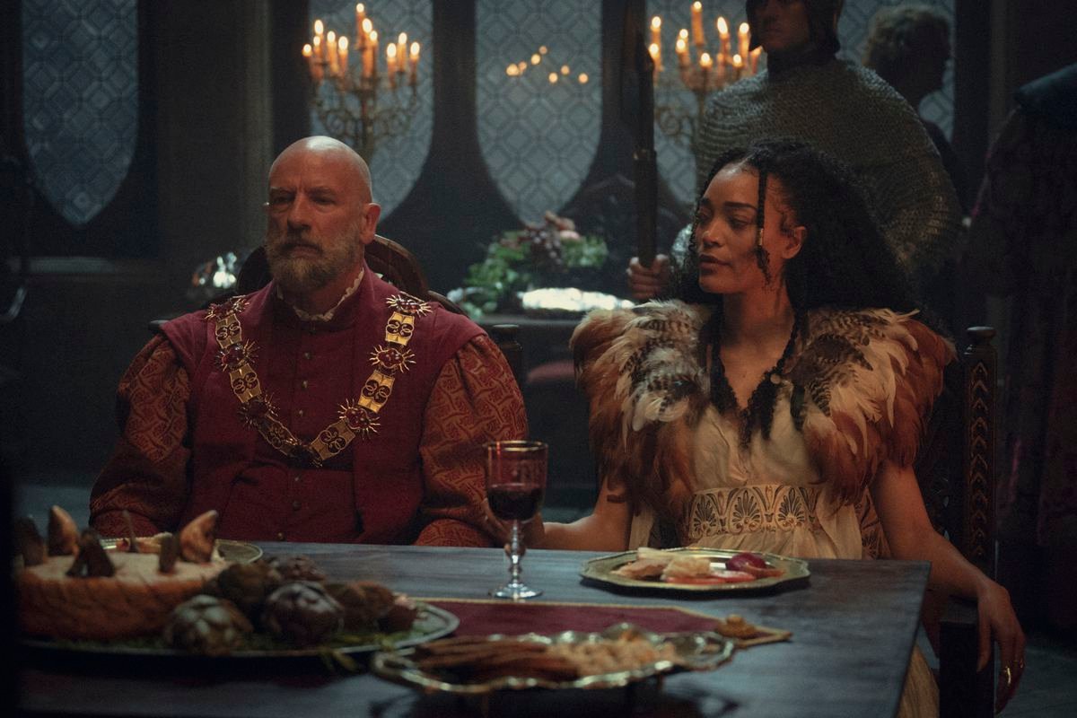 Djikstra and Philippa, two schemers, sit at a dinner table with a glass of wine between them and a spread at its center in season three of Netflix’s The Witcher