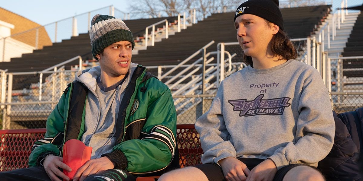 Brothers Keith and Kevin Gill (Paul Dano and Pete Davidson), in slouchy casual wear (sweatshirts, hoodies, knit hats) sit on outdoor bleachers together in Dumb Money
