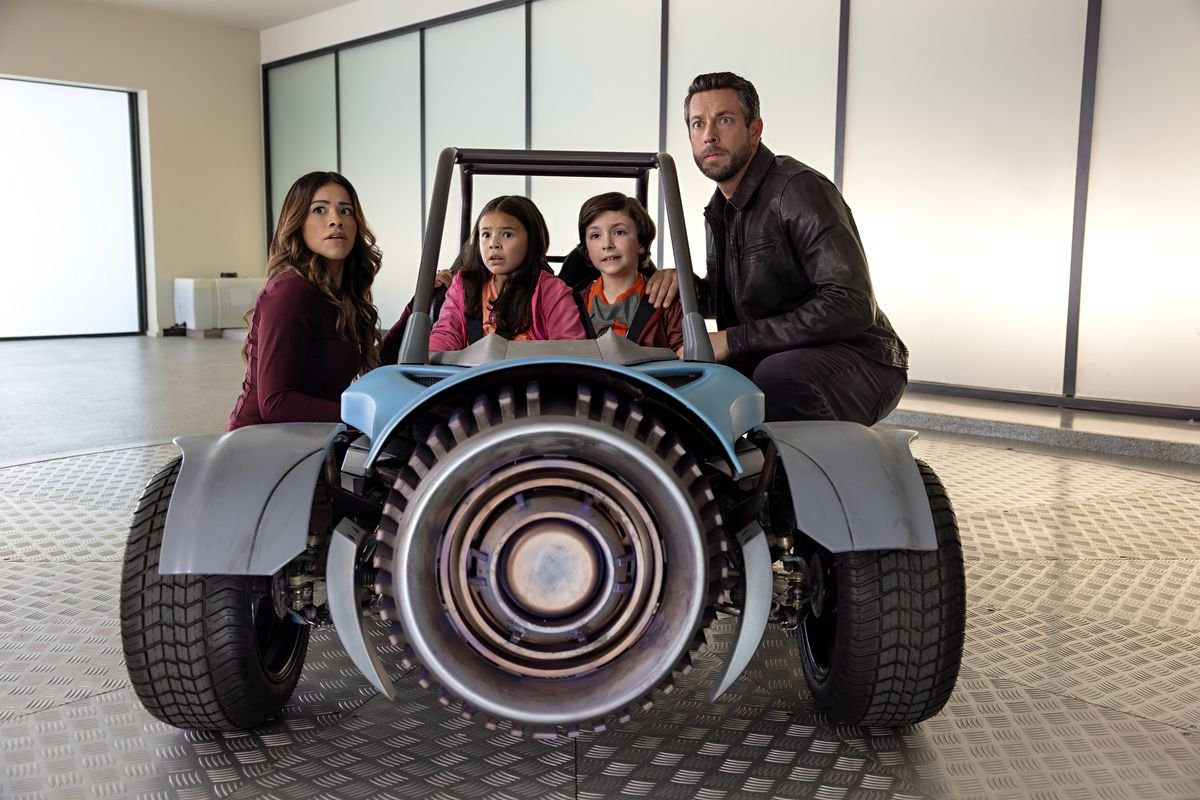 Two kids sit in a go-kart-esque vehicle. On either side of them are their parents.