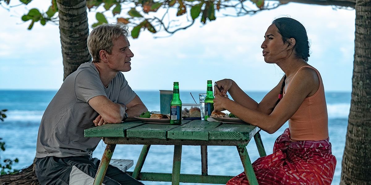 Coach Thomas Rongen (Michael Fassbender) sits at a blue picnic table across from soccer player Jaiyah Saelua (Kaimana) with the ocean as a backdrop in Next Goal Wins