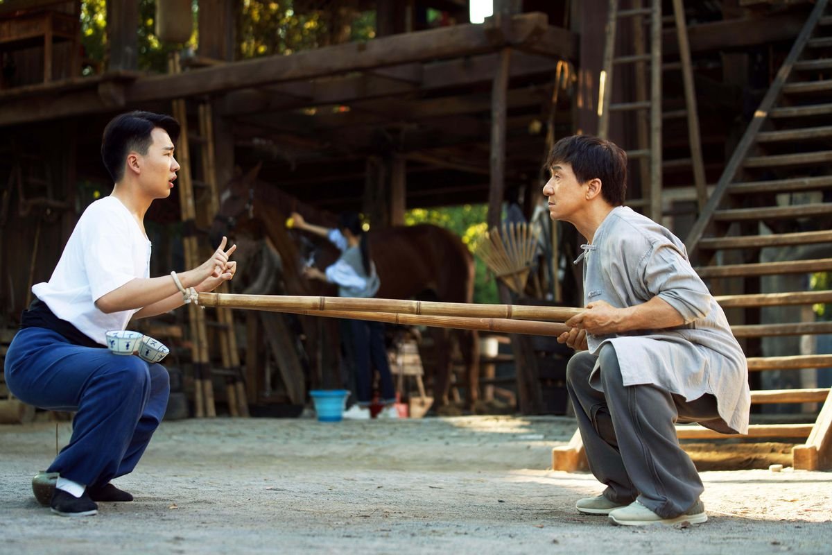 Jackie Chan and Kevin Guo perform strenuous squatting exercises as a horse stands in the background in Ride On.