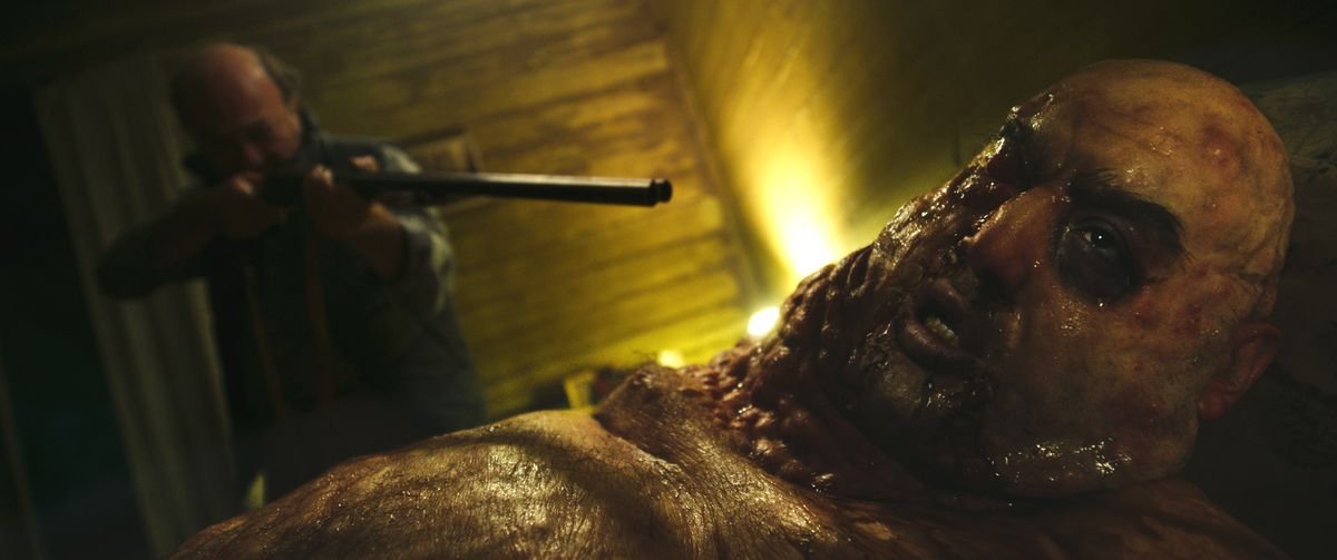 An out-of-focus man in the background of a dimly lit shack points a shotgun at the face of a figure in the foreground, a bloated, pustule-covered, bald man with sunken dark eyes, dark veins, and dark smears around his mouth in When Evil Lurks