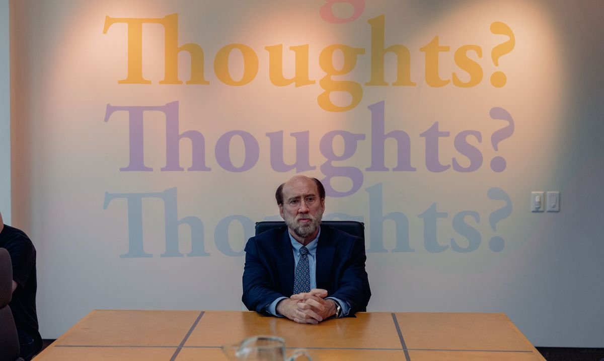Schlubby professor Paul Matthews (Nicolas Cage), in a suit and tie, sits uncomfortably at the end of a boardroom table in front of a wall with the words “Thoughts? Thoughts? Thoughts?” written on it in three different pastel shades in a scene from A24’s Dream Scenario