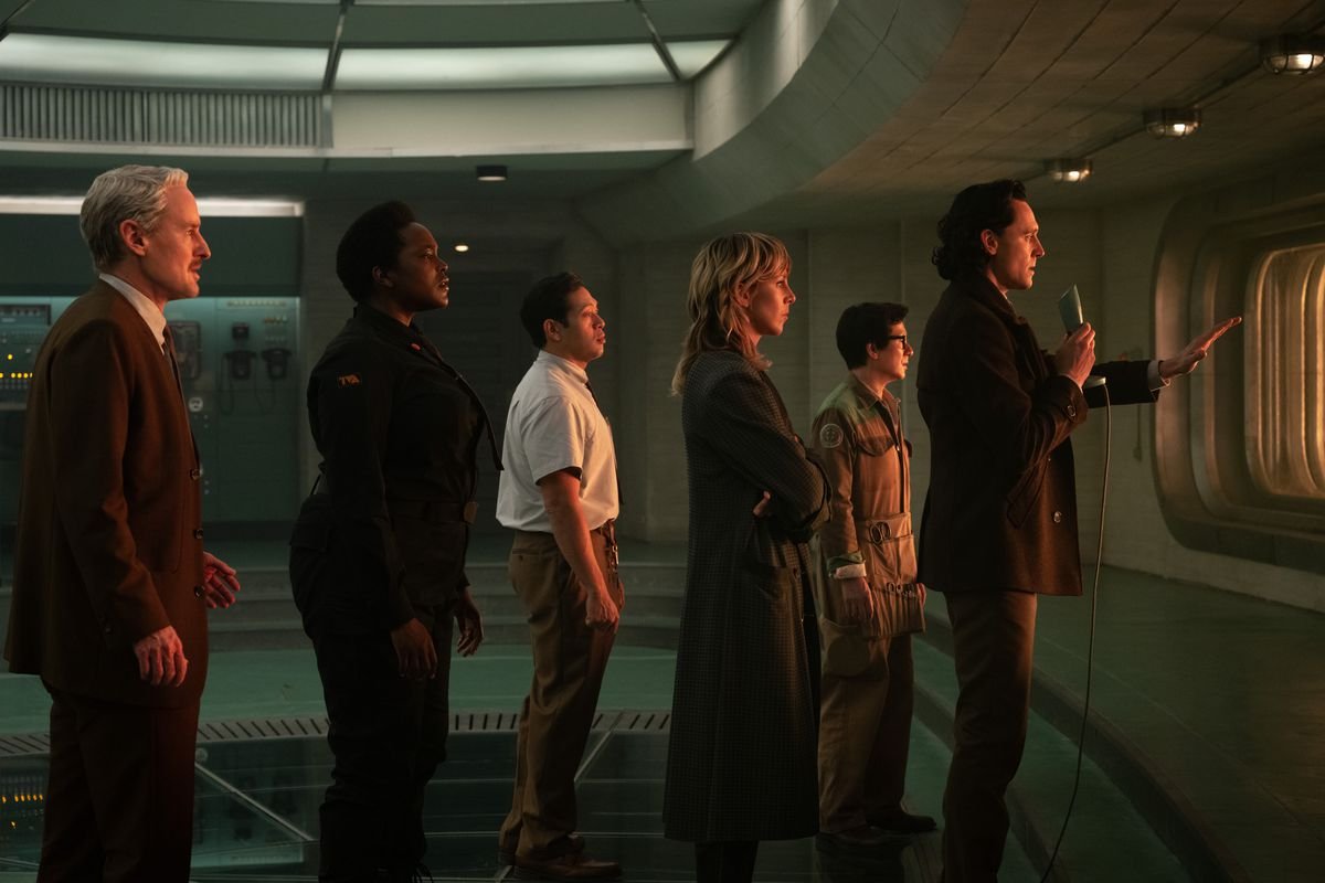 Loki (Tom Hiddleston) and the TVA crew stand looking out a window, while Loki holds a mic and raises his hand to explain something to someone on the other side of the window
