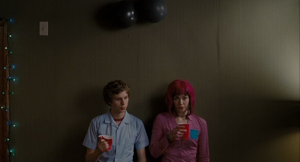 Scott Pilgrim, a young man wearing a blue buttoned up dress shirt over a graphic t-shirt stands beside Ramona Flowers, a woman with neon pink hair and a pink dress shirt, against a wall, a red party cup clutched in his hand, in Scott Pilgrim vs. the World