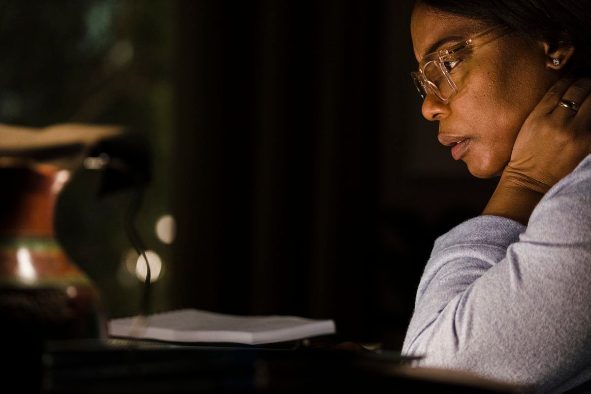 Aunjanue Ellis-Taylor, wearing spectacles and lit dimly in a close-up at nighttime, in Origin