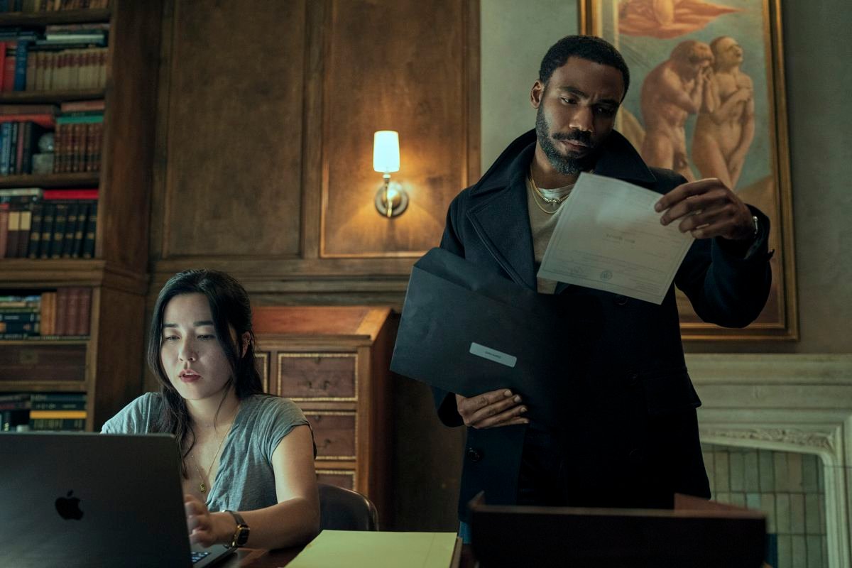 Maya Erskine looks at a laptop while Donald Glover studies a document in a fancy study in Mr. & Mrs. Smith