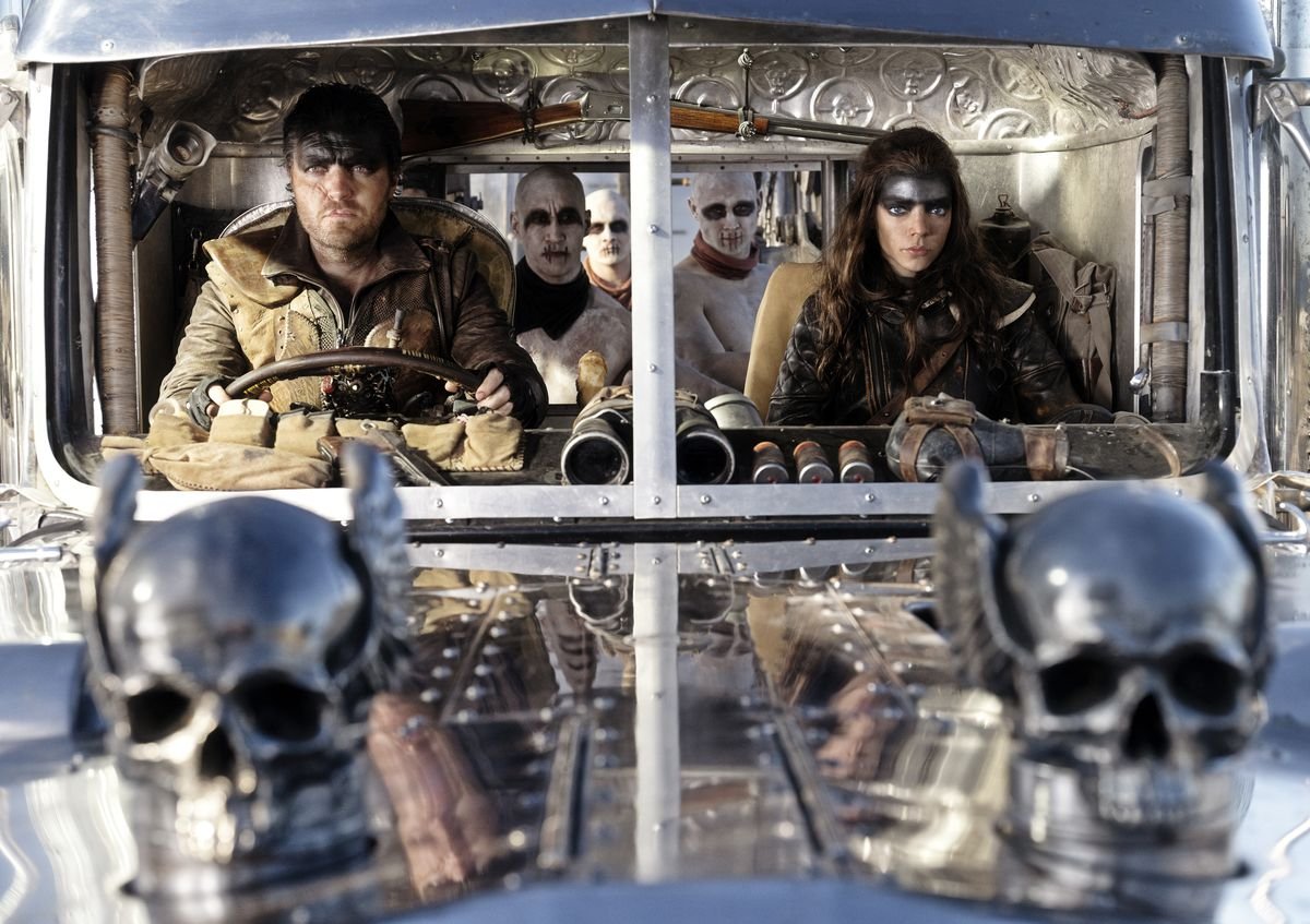 Furiosa (Anya Taylor-Joy) and Praetorian Jack (Tom Burke), two leather-clad post-apocalyptic warriors, sit in the front seats of a truck with huge silver winged skulls on the hood and a group of War Boys with shaved heads and white face paint in the back in George Miller’s Furiosa