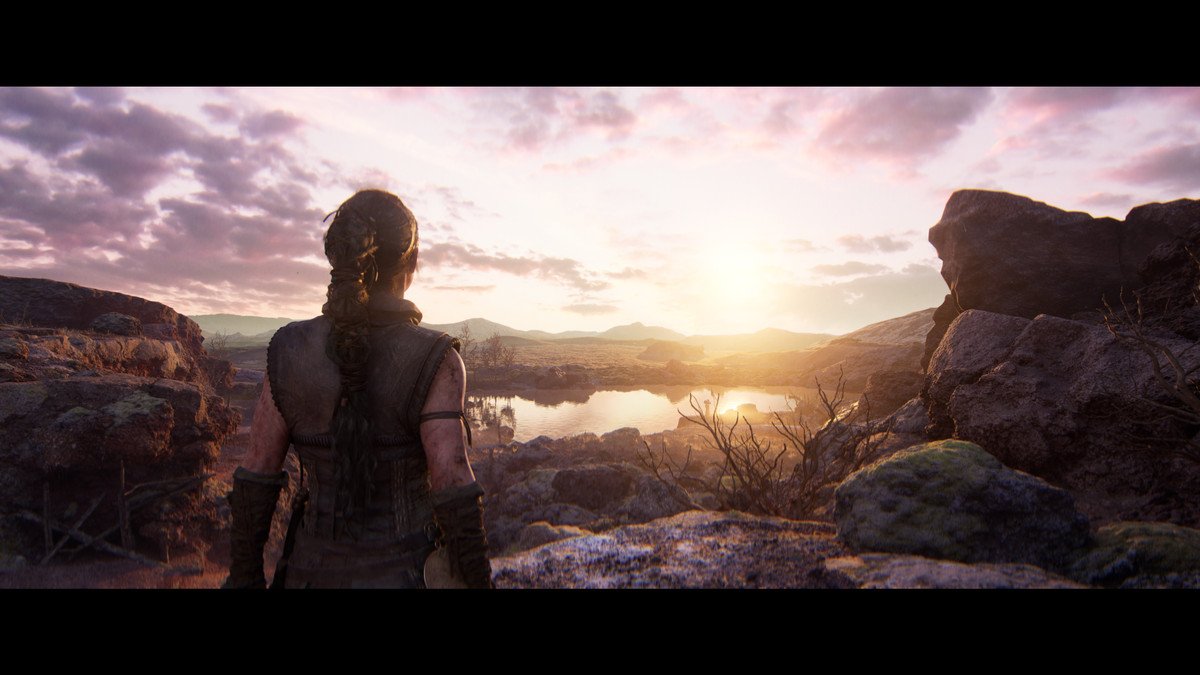 A woman with long, dark hair in a braid faces away from the camera towards a beautiful landscape in the sunrise