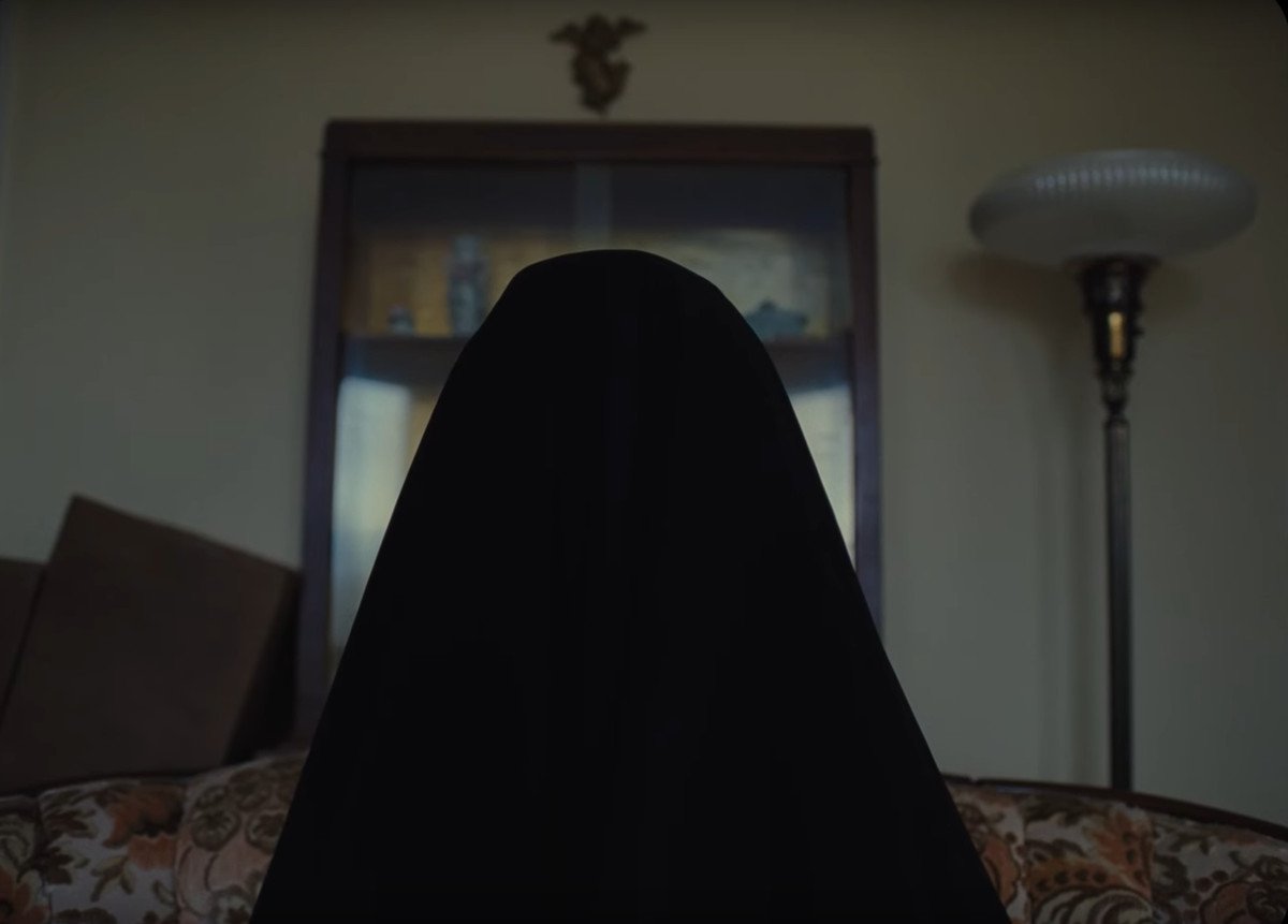 A dark figure in a ghostly habit shape fills up most of the frame while standing in front of a doorway with a cross above it in Longlegs